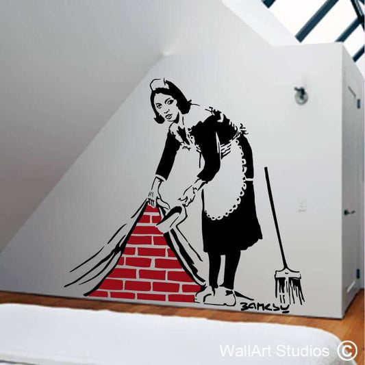 A Banksy Maid in London wall sticker depicting a woman cleaning a room against a brick wall.